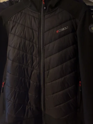 Svart vindjacka från Cobolt med huva - Snygg svart vindjacka från Cobolt med huva och hel dragkedja. Jackan har vadderade partier framtill, två sidofickor med dragkedja och diskret logga på bröstet. Materialet är en mix av softshell och quiltad polyester, perfekt för blåsiga dagar.