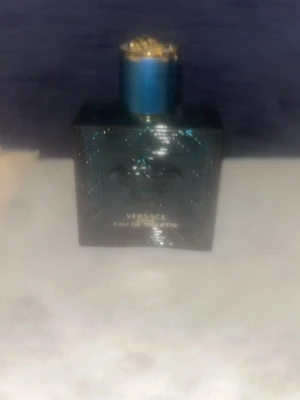 Tom Versace Eau de Toilette blå flaska - Snygg parfym från Versace i en mörkblå, fyrkantig glasflaska med gulddetaljer på korken. Flaskan har ett elegant mönster och känns riktigt lyxig. Perfekt för dig som gillar exklusiva dofter och vill ha en stilren parfymflaska i badrumshyllan.