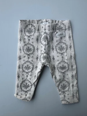 Vita leggings med blommönster Newbie - Supersöta vita leggings från Newbie med grått blommönster och rosetter. Mjukt och stretchigt material med resår i midjan för skön passform. Perfekta att matcha med en enkel topp eller body.