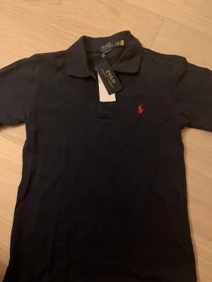 Svart t-shirt från Ralph Lauren - Klassisk svart t-shirt från Ralph Lauren med röd broderad logga på bröstet. T-shirten har korta ärmar och rund halsringning. Perfekt basplagg med stilren design.