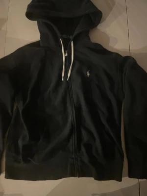 Svart hoodie från Polo Ralph Lauren - Snygg svart hoodie från Polo Ralph Lauren med dragkedja framtill och vit broderad logga på bröstet. Hoodien har vita dragsnören och är tillverkad i mjukt bomullsmaterial. Perfekt för en chill och stilren look.