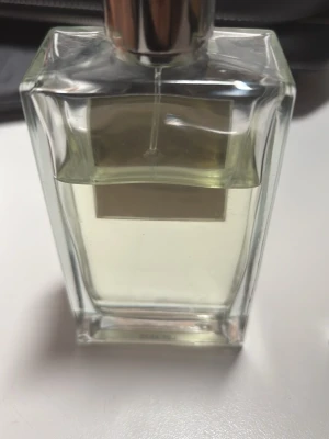 Elegant parfymflaska med silverlock - Snygg parfymflaska i genomskinligt glas med rektangulär form och stilrent silverfärgat lock. Flaskan innehåller en ljusgul vätska och har en minimalistisk etikett på framsidan. Perfekt för dig som gillar klassisk och stilren design.