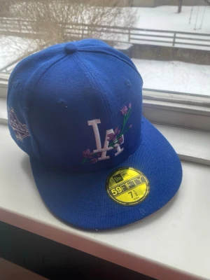 Blå LA Dodgers 59Fifty keps - Säljer en blå New Era 59Fifty keps med LA Dodgers-logga framtill och broderade blommor. På sidan finns ett 1988 World Series-märke. Klassisk flat peak och broderade ventilationshål. Perfekt för dig som gillar sportig streetstyle.