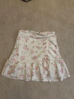 Vit blommig kortkjol med volang - Supersöt vit kortkjol med rosa blommor och gröna blad. Kjolen har volangkant nertill och snörning framtill som ger en extra detalj. Perfekt för sommarens alla dagar och lätt att matcha med en enkel topp.