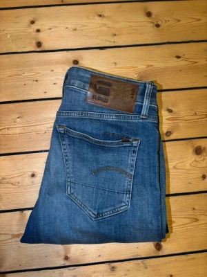 Blå jeans från G-Star RAW - Säljer ett par klassiska blå jeans från G-Star RAW med snygg tvätt och raka ben. Jeansen har fem fickor, läderpatch med logga bak och coola detaljer vid bakfickan. Perfekta för dig som gillar streetwear och avslappnad stil.