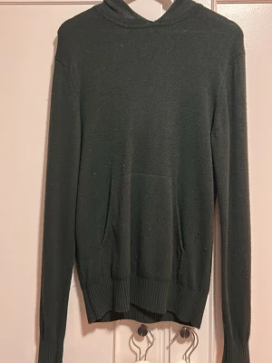 Mörkgrön hoodie med huva från Mango - En stilren mörkgrön hoodie med huva och magficka. Tillverkad i ett mjukt, tunt stickat material som känns skönt mot huden. Långa ärmar och ribbade muddar vid ärmslut och nederkant ger en snygg passform. Perfekt för en avslappnad look.