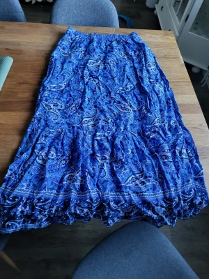 Blå långkjol med paisleymönster - Säljer en blå långkjol med paisleyinspirerat mönster i olika blå nyanser. Kjolen har resår i midjan och ett luftigt, böljande fall som ger en avslappnad och bohemisk känsla. Perfekt för dig som gillar färgstarka plagg med mycket mönster.