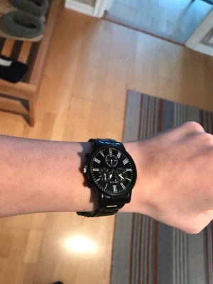 Svart klocka  - Stilren svart klocka med metallarmband och svart urtavla. Klockan har romerska siffror, flera små urtavlor och en klassisk design som passar till det mesta. Perfekt accessoar för dig som gillar en clean och modern look.