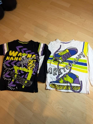 Två långärmade tröjor från KappAhl - Säljer två coola långärmade tröjor från KappAhl. Ena är svart med lila, gula och vita detaljer samt skateboard-tryck och texten 'WANNA HANG OUT'. Den andra är vit med färgglada sneakers och texten 'ARE YOU READY?'. Perfekta för dig som gillar streetstyle.