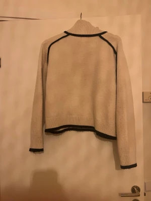 Beige polotröja med svarta detaljer - Stickad beige polotröja med svarta kontrastsömmar och ribbade muddar. Tröjan har en hög krage och är något croppad i modellen. Perfekt för dig som gillar en enkel men ändå unik look.