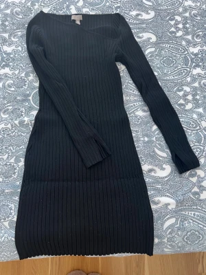 Svart ribbad långärmad klänning - Snygg svart ribbad klänning med lång ärm och kroppsnära passform. Klänningen har en enkel, stilren design och är tillverkad i ett stretchigt material som formar sig efter kroppen. Perfekt för dig som gillar minimalistisk stil.