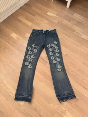 Blå jeans med tryck från Gallery dept - Unika blå jeans från Gallery dept med grått G-tryck längs båda benen framtill. Jeansen har fem fickor, raka ben och råa, fransiga benslut för en avslappnad vibe. Tillverkade i klassisk denim med slitningar för extra edge.