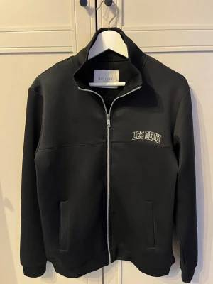 Svart jacka med dragkedja från Les Deux - Stilren svart jacka från Les Deux med hög krage och hel dragkedja framtill. Jackan har två fickor och diskret broderad logga på bröstet. Perfekt för dig som gillar minimalistisk design och vill ha en enkel men snygg jacka.