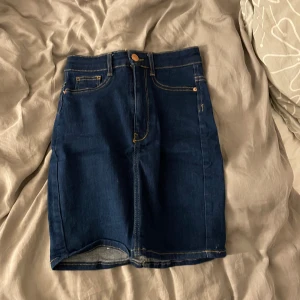 Mörkblå jeanskjol med fickor - Snygg mörkblå jeanskjol med klassiska gula sömmar och fem fickor. Kjolen har en kort passform och stängs med knapp och dragkedja framtill. Perfekt att matcha med en enkel t-shirt eller hoodie för en avslappnad look.