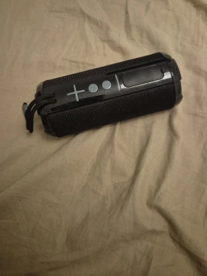 Svart bärbar högtalare med tygfront - Snygg svart bärbar högtalare med robust tygfront och tydliga knappar för volym och uppspelning. Cylindrisk form med praktisk snodd för enkel transport. Perfekt för att ta med sig musiken överallt.