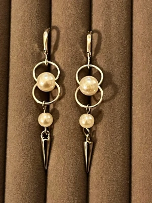 Silvriga örhängen med pärlor och spikes - Säljer ett par statement örhängen i silverfärg med två vita pärlor på varje örhänge. Designen har dubbla ringar och en cool metallspets längst ner. Perfekt för dig som gillar en mix av klassiskt och edgy stil.