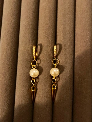 Guldörhängen med pärlor och spikes - Snygga örhängen i guldfärg med vita pärlor och spetsiga hängande spikes. Örhängena har små ringar och detaljerad yta på själva ringen. Perfekt för dig som gillar en mix av klassiskt och edgy stil.