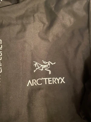 Svart vindjacka från Arc'teryx - Snygg svart vindjacka från Arc'teryx med huva och dragkedja framtill. Jackan har logga på bröstet och elastiska muddar vid ärmsluten. Tillverkad i lätt och slitstarkt material, perfekt för blåsiga dagar och outdoor-äventyr.