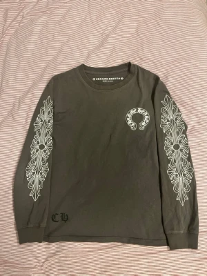 Grå långärmad topp från Chrome Hearts - Säljer en grå långärmad topp från Chrome Hearts med vita grafiska tryck längs båda ärmarna och logga på bröstet samt ryggen. Tröjan har rund halsringning och är tillverkad i mjuk bomull. Perfekt för dig som gillar streetwear och unika detaljer.
