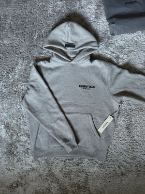 FOG Essentials Hoodie - Elegant hoodie från Essentials Fear of God i grå färg – minimalistisk design med ikonisk logga på bröstet. En stilren hoodie som kombinerar komfort med modern streetwear-estetik. ♦️ Storlek XS – avslappnad  passform ⚜️ Helt ny med tagg ♦️ Mjuk & högkvalitativ bomullsblandning 📦 Fraktas snabbt & trygg affär