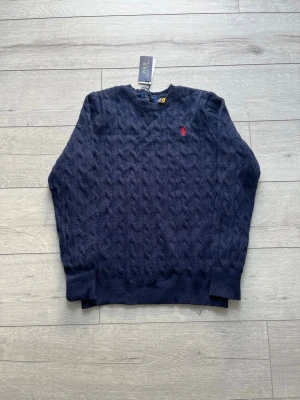 Mörkblå kabelstickad tröja Polo Ralph Lauren - Mörkblå kabelstickad tröja från Polo Ralph Lauren med rund halsringning och röd broderad logga på bröstet. Tröjan har ribbade muddar vid ärmslut och nederkant. Perfekt för dig som gillar klassisk och stilren design.
