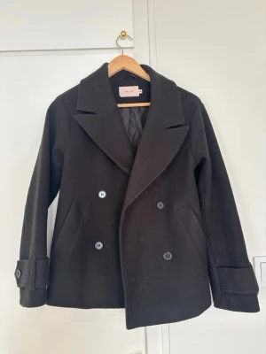 Svart kappa från Nelly - Stilren svart kappa från Nelly med dubbelknäppning och breda slag. Jackan har två sidofickor och dekorativa knappar vid ärmsluten. Tillverkad i ett tjockare material som ger en klassisk look och passar perfekt till höst och vinter.