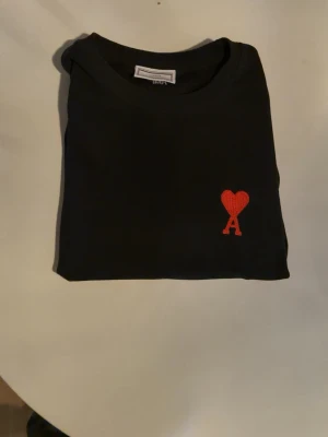 Svart sweatshirt från Ami Paris - Säljer en svart sweatshirt från Ami Paris med det ikoniska röda hjärtat och bokstaven A broderat på bröstet. Tröjan har rund halsringning och långa ärmar. Perfekt för en clean och stilren look.