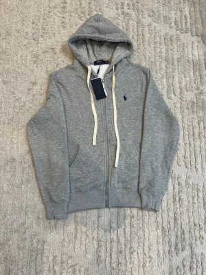 Grå hoodie från Polo Ralph Lauren - Snygg grå zip-hoodie från Polo Ralph Lauren med vit dragsko och klassisk blå logga på bröstet. Tröjan har fickor framtill, ribbade muddar och mjukt material som känns skönt mot huden. Perfekt för en avslappnad och stilren look.