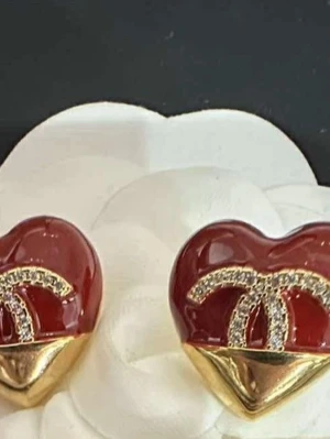 Röda hjärtformade örhängen Nyvara  - Snygga örhängen  i form av röda hjärtan med gulddetaljer och glittrande stenar på loggan. Örhängena har en lyxig känsla med sin kombination av rött, guld och bling. Perfekta för att lyfta din stil med en ikonisk touch.