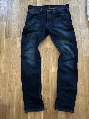 Mörkblå jeans från Lee  - Säljer ett par mörkblå jeans från Lee i slim modell. Jeansen har fem fickor, knappgylf och subtila slitningar för en snygg look. Tillverkade i kraftig denim som ger en schysst passform och stilren vibe.