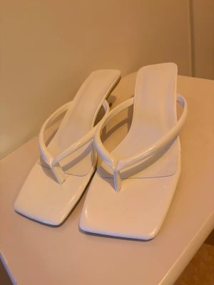 Vita trendiga sandaletter med klack - Säljer ett par helt oanvända  vita sandaletter med fyrkantig tå och smala remmar. De har en låg klack och modern design, perfekta för dig som gillar stilrena och minimalistiska skor. Materialet ser ut att vara konstläder med glansig finish.