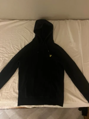 Svart hoodie från Lyle & Scott - Säljer en svart hoodie från Lyle & Scott med klassisk gul logga på bröstet. Hoodien har huva med dragsko och en stor magficka framtill. Tillverkad i mjukt bomullsmaterial och har ribbade muddar vid ärmslut och nederkant. Den har små hål på vänster arm bak, men här troligen att laga med nål och tråd, 7/10 i skick.