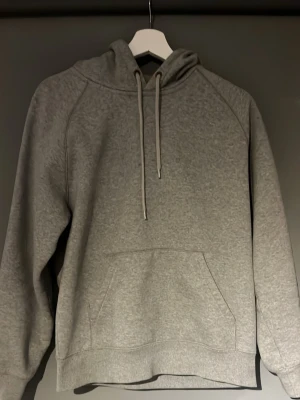 Grå hoodie från Carhartt - Säljer en klassisk grå hoodie från Carhartt med huva och dragsnören. Hoodien har en stor magficka och ribbade muddar vid ärmslut och nederkant. Carhartt-loggan i gult sitter på ärmen. Perfekt för en chill och avslappnad stil.