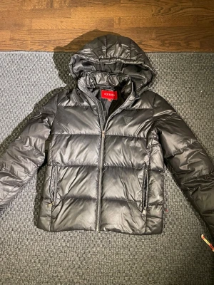Svart pufferjacka från Guess - Snygg svart pufferjacka från Guess med huva och dragkedja framtill. Jackan har två fickor med dragkedja och Guess-logga på bröstet. Tillverkad i glansigt material som ger en modern look. Perfekt för kalla vinterdagar. Passar som s mer