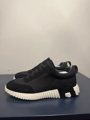 Svarta H sneakers med vit sula - Stilrena svarta sneakers med chunky vit sula och coola detaljer på sidan. Ovandelen är i textil och mockaimitation, med svarta skosnören och en låg profil. Perfekta för dig som gillar en modern och sportig look. Helt oanvända