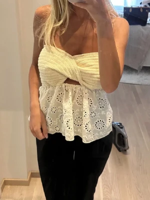 Vit axelbandslös spets topp - Supersöt axelbandslös topp i vitt med virkad överdel och broderad spets nedtill. Toppen har bandeau-form och en liten cut-out framtill. Perfekt för sommarens alla tillfällen!