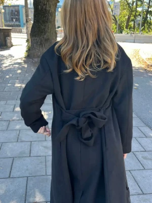 Svart lång kappa med knytskärp - Stilren svart kappa med knytskärp i midjan och klassisk krage. Lång modell som ger en snygg siluett. Perfekt för dig som gillar en enkel men trendig look. Kappan har raka ärmar.