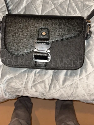 Svart axelväska från Dior - Säljer en svart axelväska från Dior med monogrammönster och detaljer i läder. Väskan har en unik metallspänne framtill och justerbar axelrem. Perfekt för dig som vill ha en stilren och trendig väska med lyxig känsla.