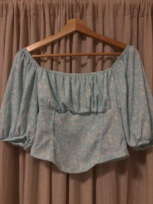 Blommig offshoulder topp Gina Tricot - Supersöt ljusblå offshoulder topp från Gina Tricot med puffiga trekvartsärmar och vitt blommönster. Toppen har en croppad passform.