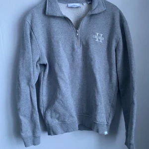 Grå halvzip sweatshirt Les Deux - Grå sweatshirt från Les Deux med halv dragkedja och ståkrage. Tröjan har broderad logga på bröstet och är fodrad med mjukt teddyfoder på insidan. Ribbstickade muddar vid ärmslut och nederkant. Perfekt för lager-på-lager-stil.