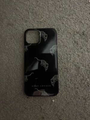 Svart mobilskal med leoparder iPhone 13 - Snyggt svart mobilskal från Roar Sweden med glittrande leopardhuvuden i silver. Skalets yta är blank och har ett stilrent tryck. Passar perfekt för dig som vill ha ett unikt och trendigt skydd till din mobil.