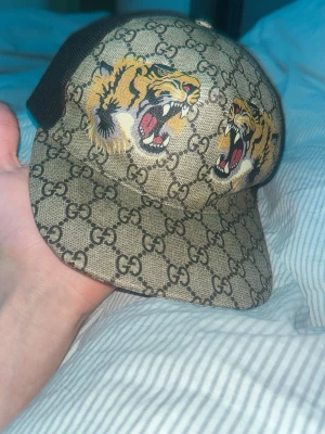 Gucci keps med tigertryck - Snygg Gucci keps med klassiskt GG-mönster i beige och brunt, prydd med två tigrar framtill. Baksidan är svart mesh med justerbart band i grönt och rött. Perfekt för dig som vill sticka ut med en exklusiv accessoar.