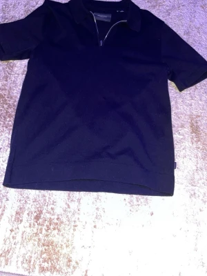 Svart kortärmad t-shirt med half zip - Stilren svart t-shirt från Weekday med korta ärmar och krage. Tröjan har en snygg half zip-dragkedja framtill som ger en modern touch. Perfekt till jeans eller kjol för en clean look.