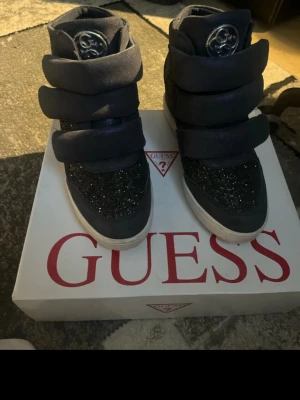 Svarta höga sneakers från Guess - Snygga svarta sneakers från Guess med högt skaft och tre breda kardborreband. Skorna har glittrande detaljer på ovandelen och en vit sula. Guess-logga framtill och på insidan. Perfekta för dig som vill sticka ut med stilrena detaljer.