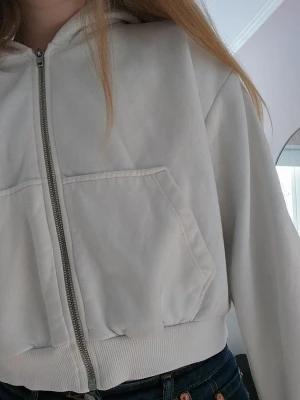 Vit zip hoodie med huva - Snygg vit hoodie med dragkedja och huva. Modellen är croppad med breda muddar och stora fickor framtill. Perfekt för en chill och avslappnad stil. Materialet känns mjukt och skönt mot huden.