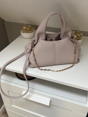 Ljuslila handväska med guldkedja - Trendig ljuslila handväska i mjukt, strukturerat konstläder. Väskan har både ett kort handtag och en längre axelrem samt en snygg guldfärgad kedjedetalj. Stängs med magnetknapp och har en stilren, modern form som passar till många outfits.