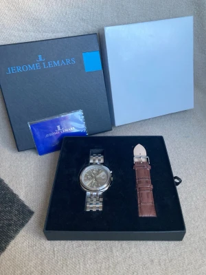 Jerome Lemars klocka med extra armband - Stilren klocka från Jerome Lemars med silverfärgat metallarmband och mörk urtavla. Medföljer ett extra brunt armband i skinn för att enkelt byta stil. Klockan levereras i originalbox med certifikat.