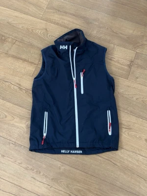 Helly Hansen väst - Sportig marinblå väst från Helly Hansen med vita dragkedjor och röda detaljer. Västen har hög krage, tre fickor med dragkedja och logga både fram och bak i nacken. Perfekt för lager-på-lager och outdoor-looks.