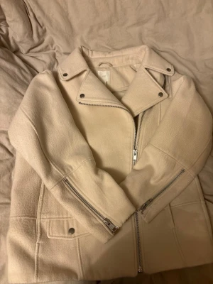Krämvit ullkappa från H&M - Snygg krämvit kappa från H&M med bred krage och silvriga dragkedjor. Kappan har en rak passform, stora fickor framtill och coola detaljer vid ärmsluten. Materialet ser ut att vara ullblandning, perfekt för kyligare dagar. Enkel men trendig design som lyfter din outfit.