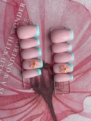 Press-on naglar rosa med blå tip - Snygga press-on naglar i ljusrosa med ljusblå fransk tip och 3D-blommor på två naglar. Naglarna har en oval form och en glansig finish.